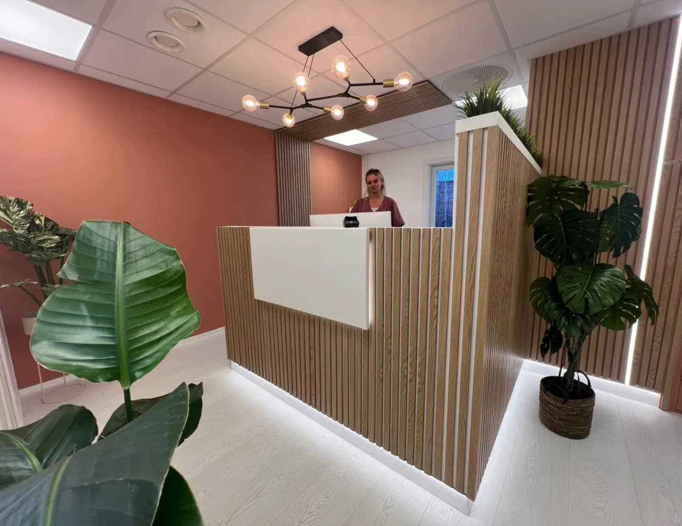 Her ser vi resepsjonsområdet ved Godtanna Løten Tannlegesenter, med en kvinne som jobber bak en stilren skranke med trepanel. Rommet er dekorert med grønne planter som gir et friskt preg.