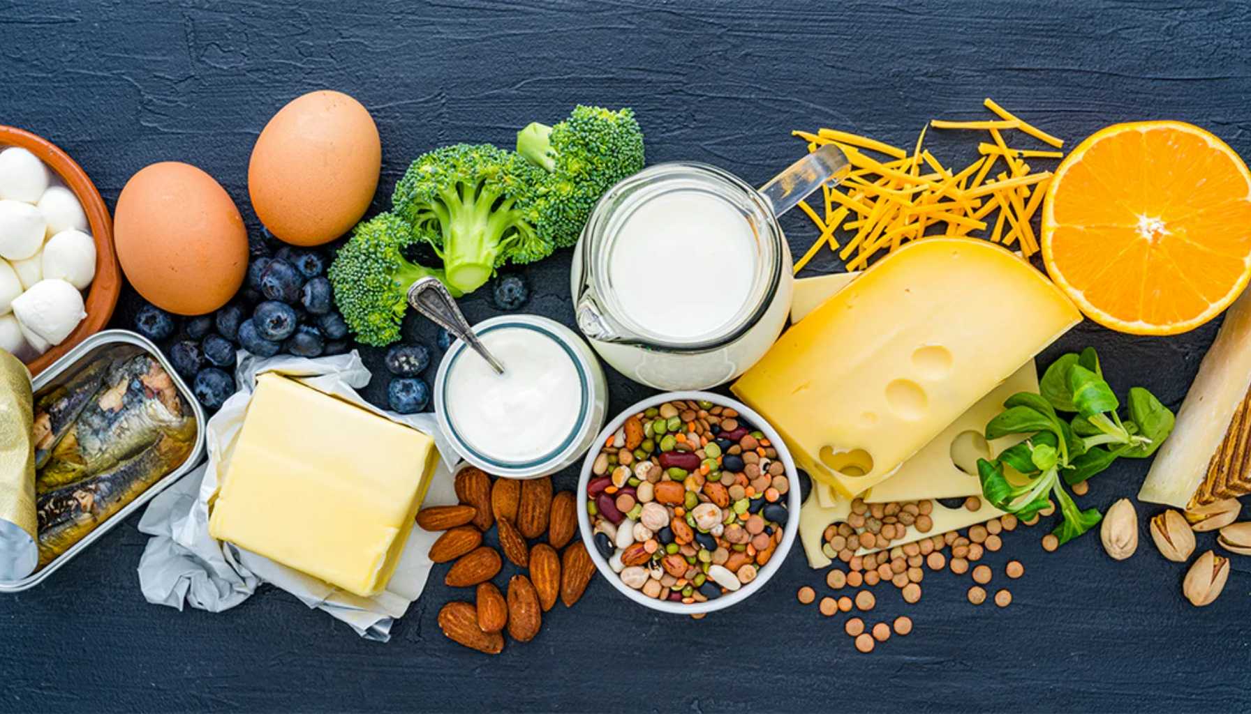 Et utvalg av kalsiumrike matvarer, inkludert ost, melk, nøtter, belgfrukter, egg, fisk, brokkoli og appelsin.