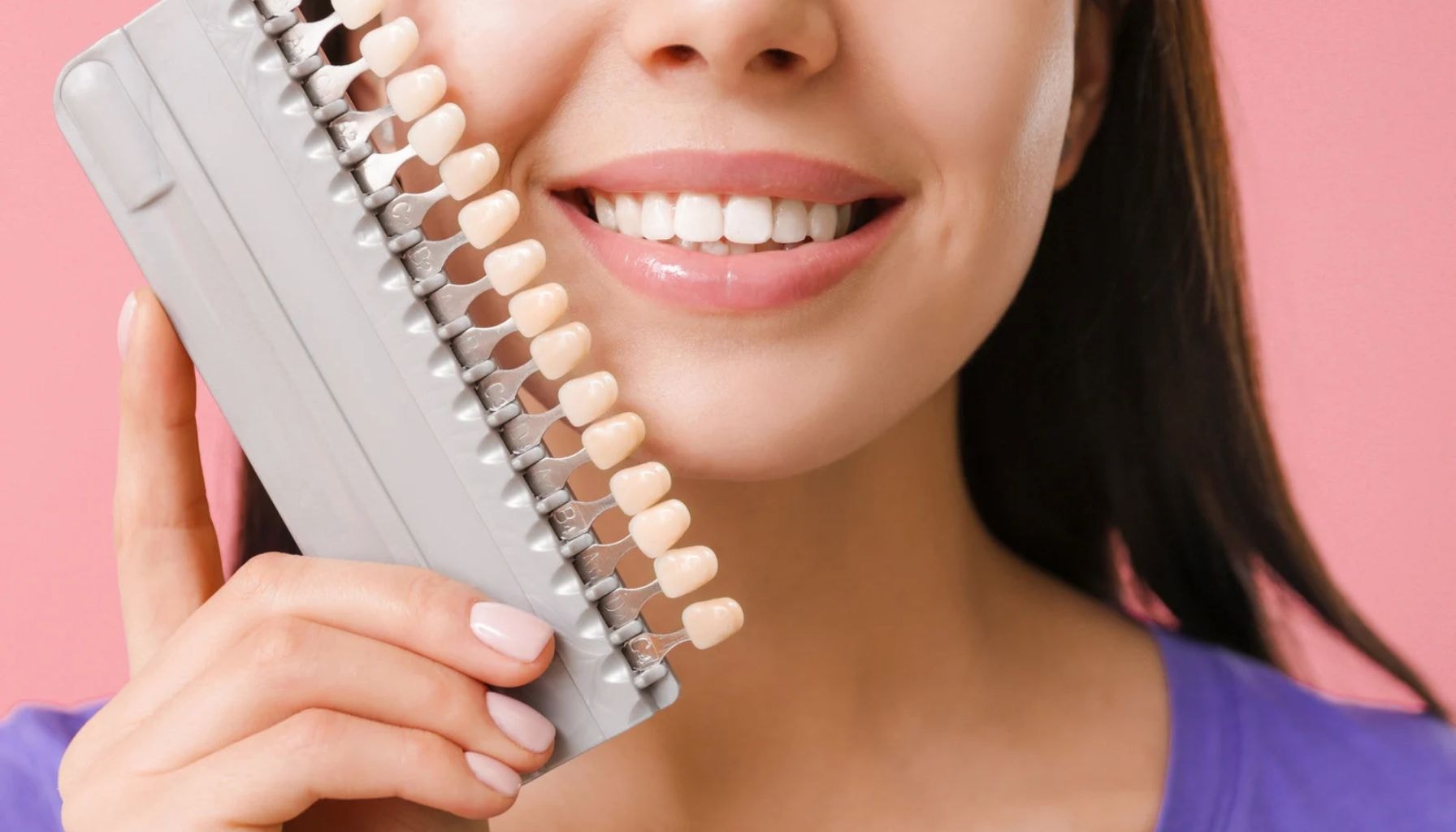 En kvinne smiler og holder en tannfargeskala ved ansiktet for å sammenligne nyanser av tannfarge mot sine egne tenner.