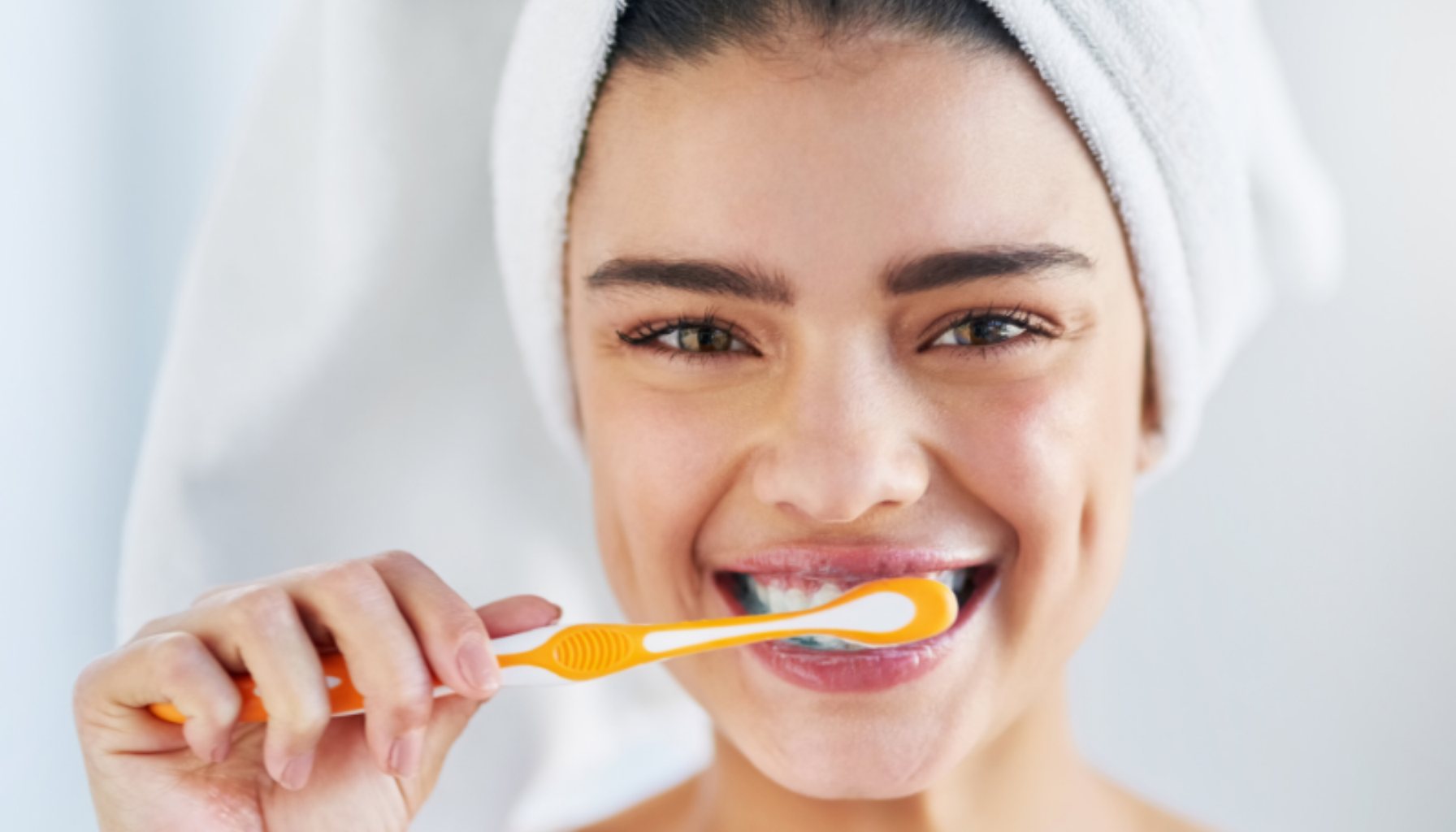 Kvinne som pusser tennene sine med en oransje tannbørste, smilende og i et håndkle på hodet