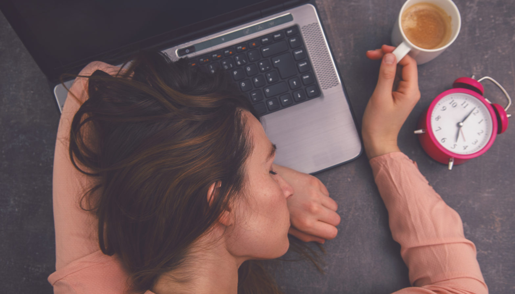 En trøtt kvinne sover med hodet på en bærbar PC, mens hun holder en kaffekopp. En vekkerklokke viser tiden på bordet. graden av symptomer