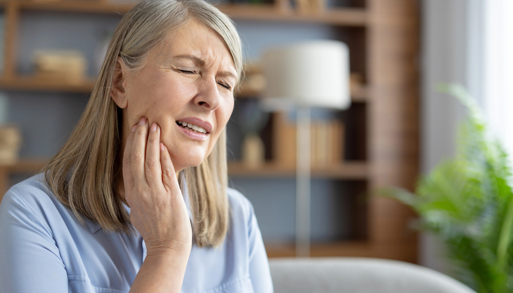 Kvinne med tannverk holder hånden mot kinnet og viser smerte, sittende i en stue med uskarp bakgrunn og planter."