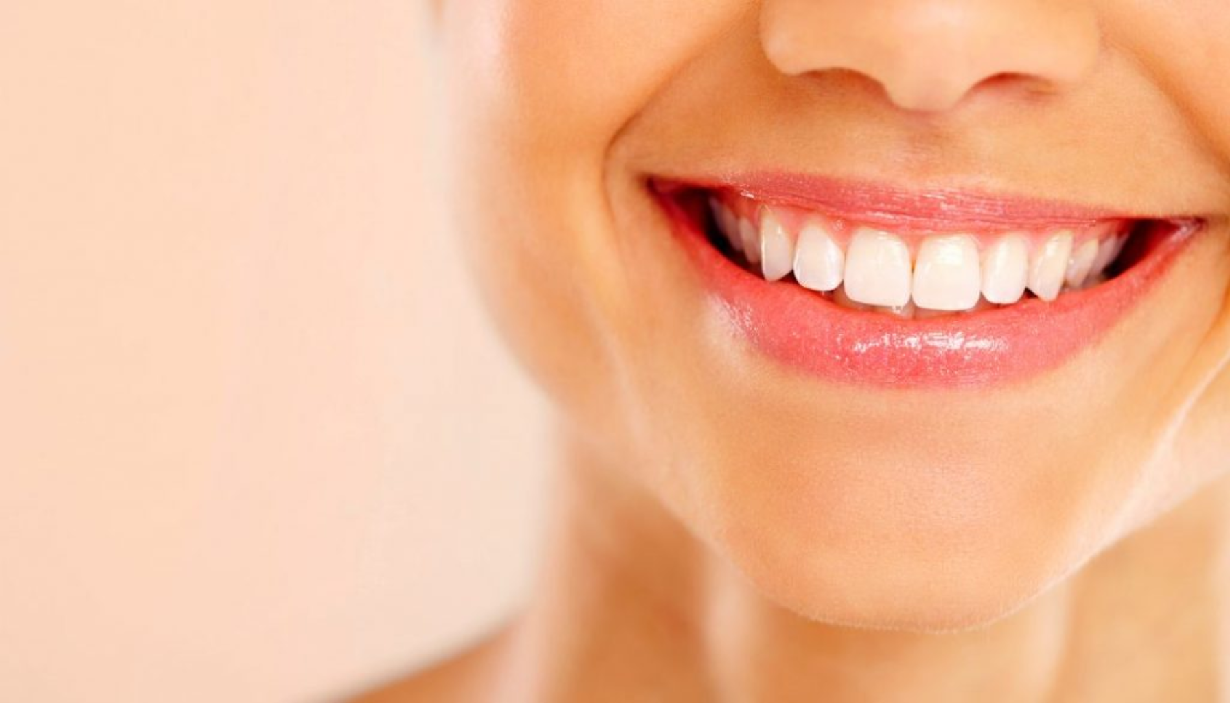 Et nærbilde av en smilende person med hvite tenner og glansfulle lepper mot en myk beige bakgrunn.