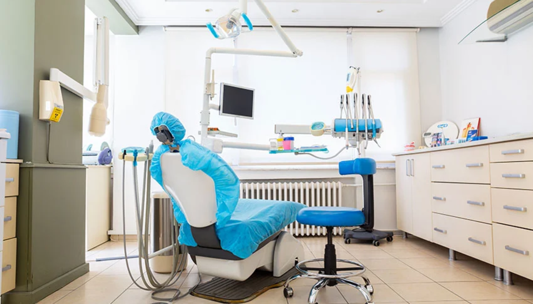 En moderne tannlegeklinikk med en tannlegestol dekket med blå beskyttelse, tannlegeutstyr, en skjerm og oppbevaringsskap i et lyst rom.