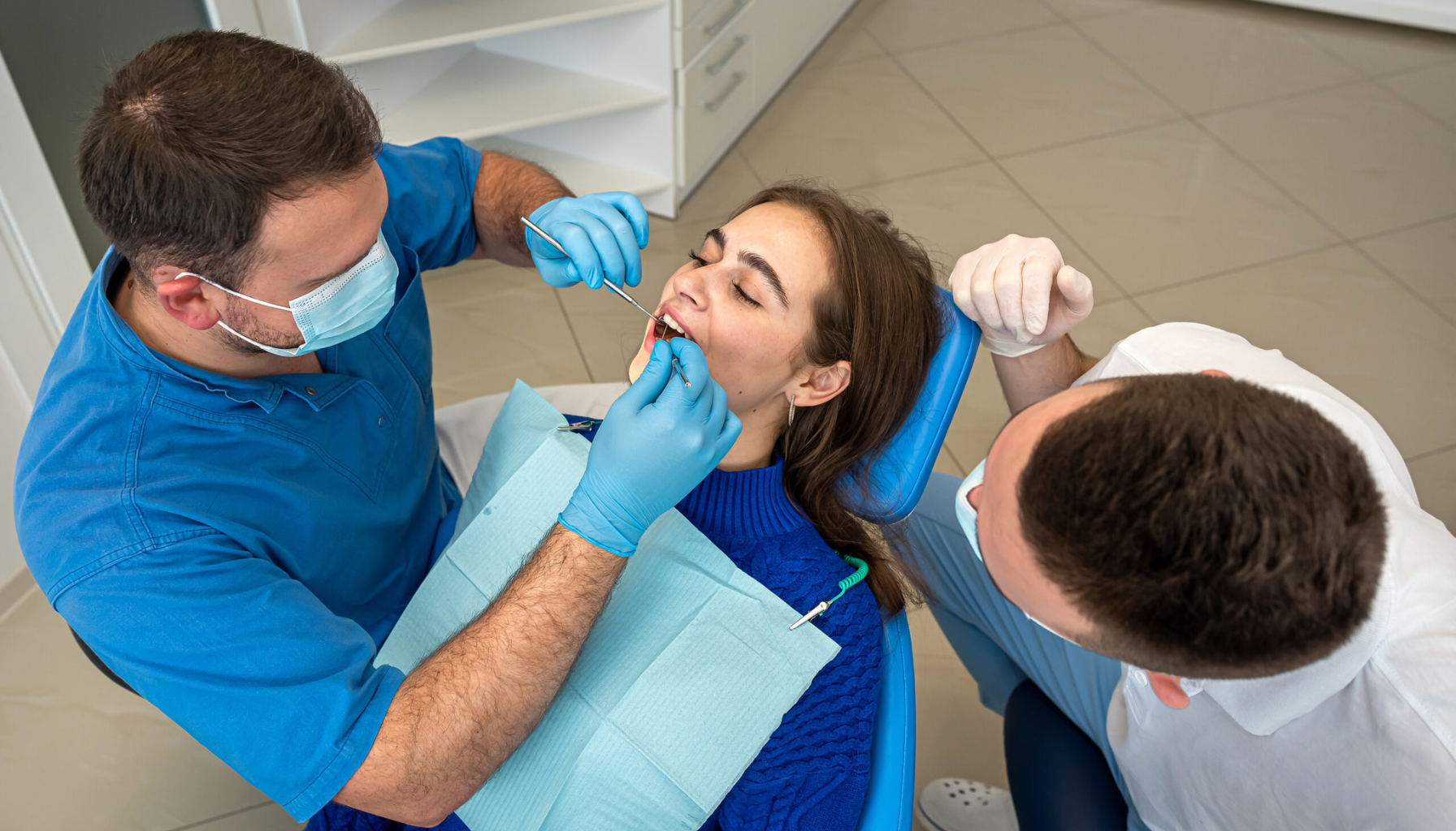 Bildet viser en kvinne som får en tannlegeundersøkelse av to tannleger i en moderne tannklinikk. Tannlegene bruker hansker og munnbind, mens pasienten ligger avslappet i en tannlegestol iført en blå genser. Det sterile og rene miljøet understreker profesjonaliteten i tannbehandlingen.