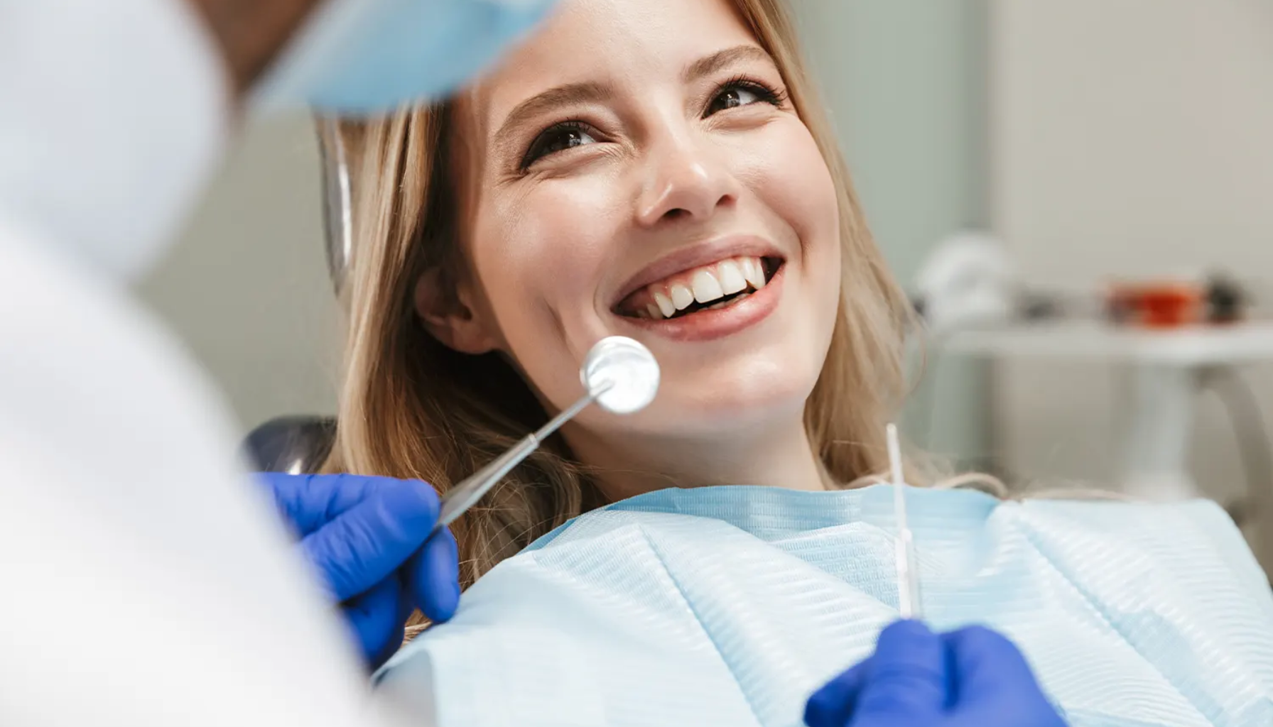 Smilende kvinnelig pasient under en tannlegeundersøkelse mens tannlegen i hansker holder et speil og en sonde.