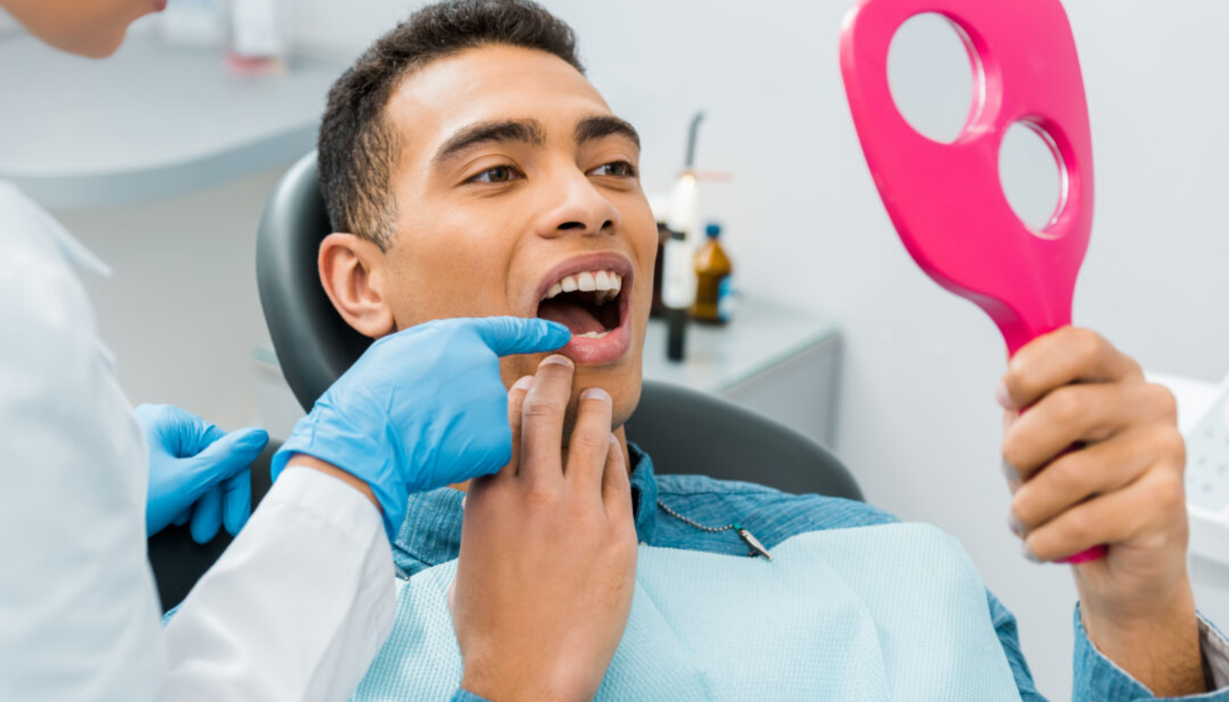 Pasient i tannlegestol ser på tennene sine i et håndspeil mens tannlegen med hansker peker på et område i munnen under undersøkelsen.