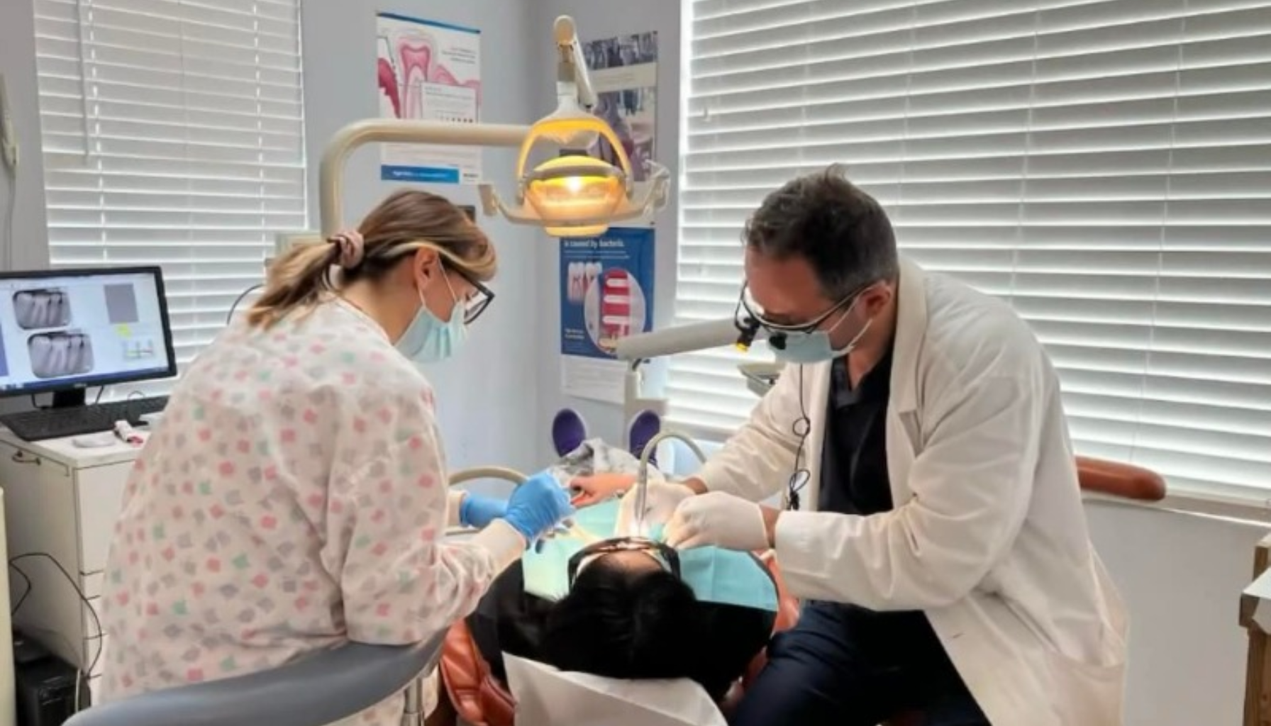 Tannlege og assistent med munnbind og hansker utfører en tannbehandling på pasient i tannlegestol under sterkt lys.