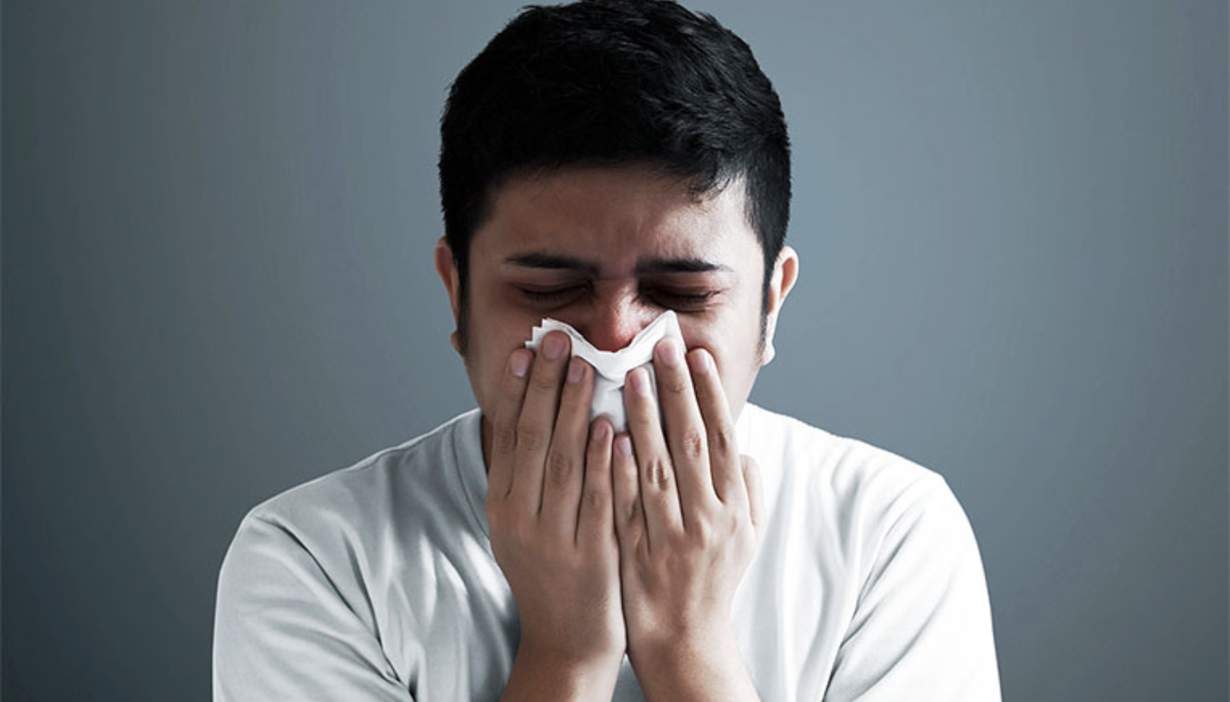 Mann med hvit t-skjorte tørker eller snyter nesen med et papir, tydelig plaget av forkjølelse eller allergi.