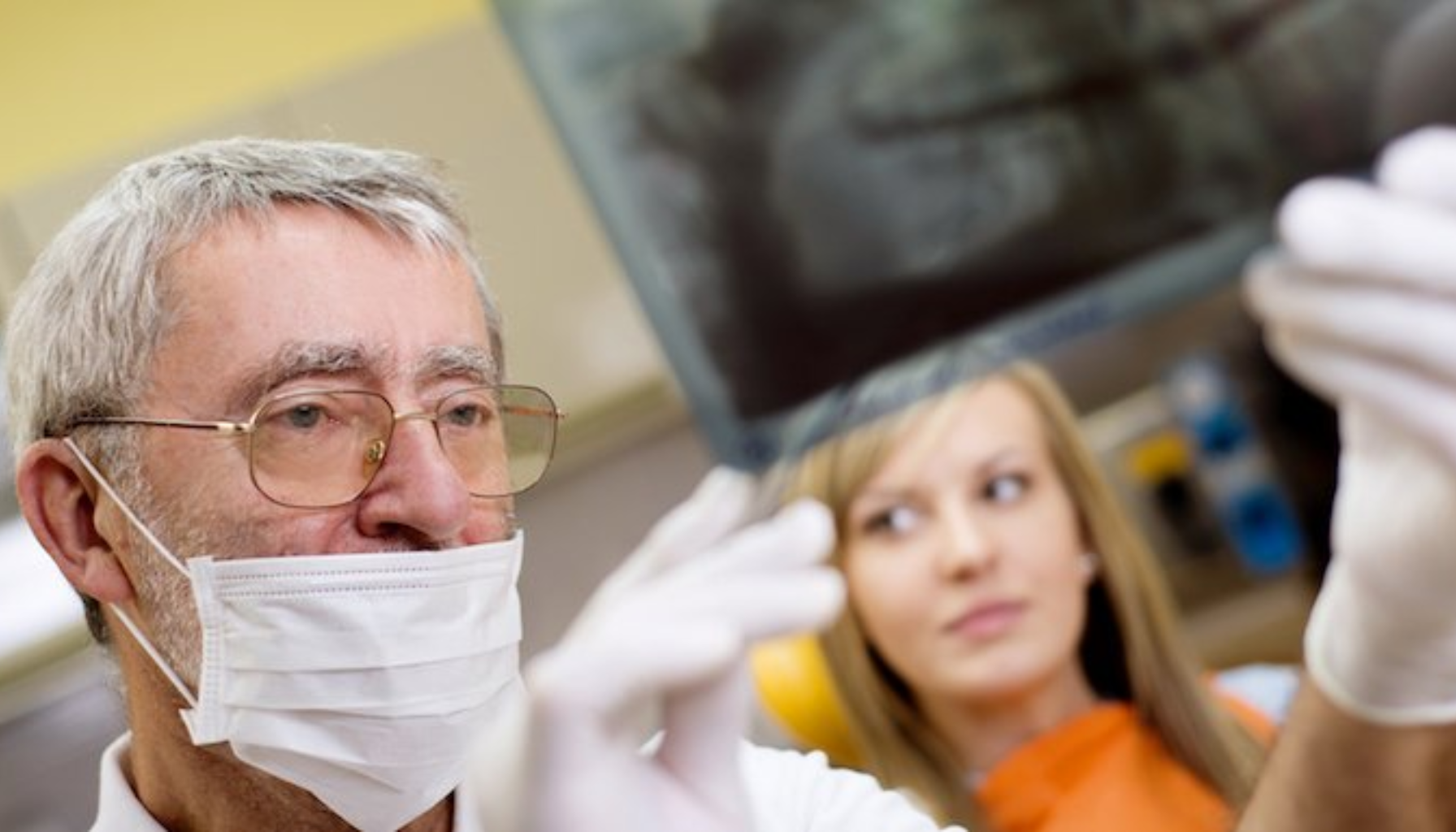 Bildet viser en tannlege som undersøker et tannrøntgenbilde mens en kvinnelig pasient sitter i bakgrunnen. Tannlegen har på seg maske og hansker, noe som tyder på at han er i ferd med å analysere bildet før eller etter en tannbehandling.