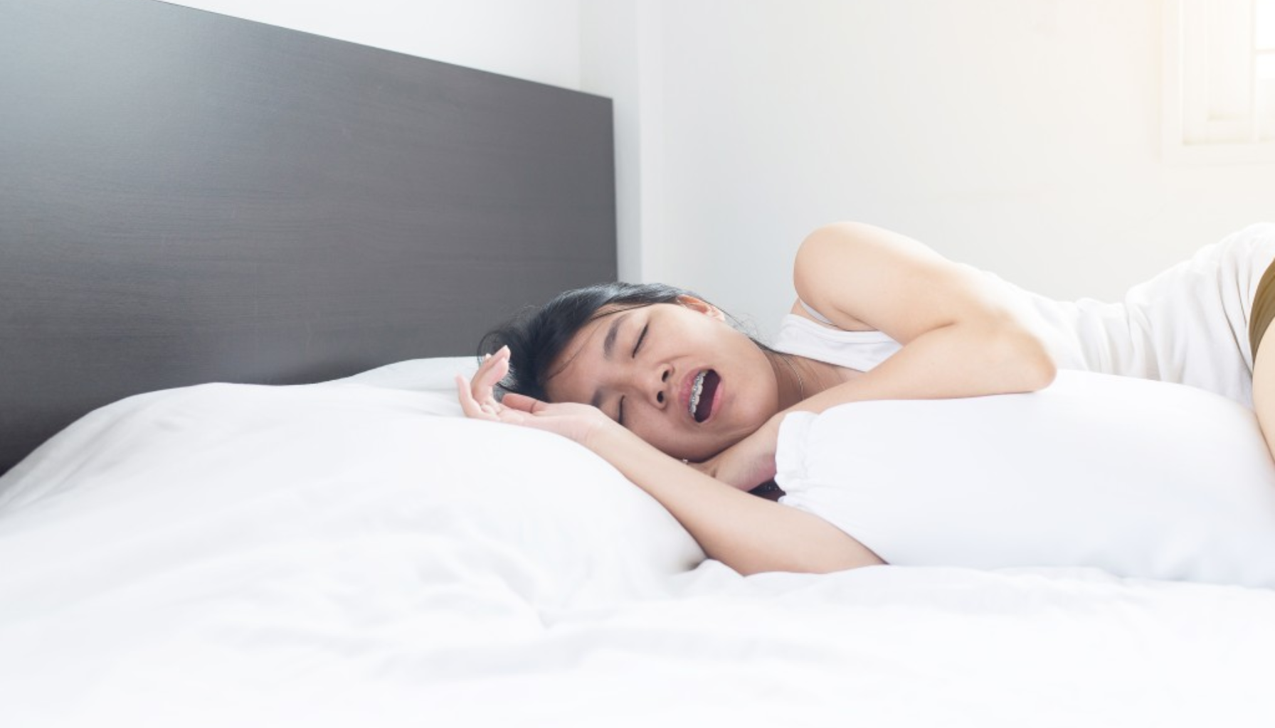 Kvinne sover på siden i en seng med hvitt sengetøy, munnen åpen, ser ut til å snorke eller puste tungt under søvn.