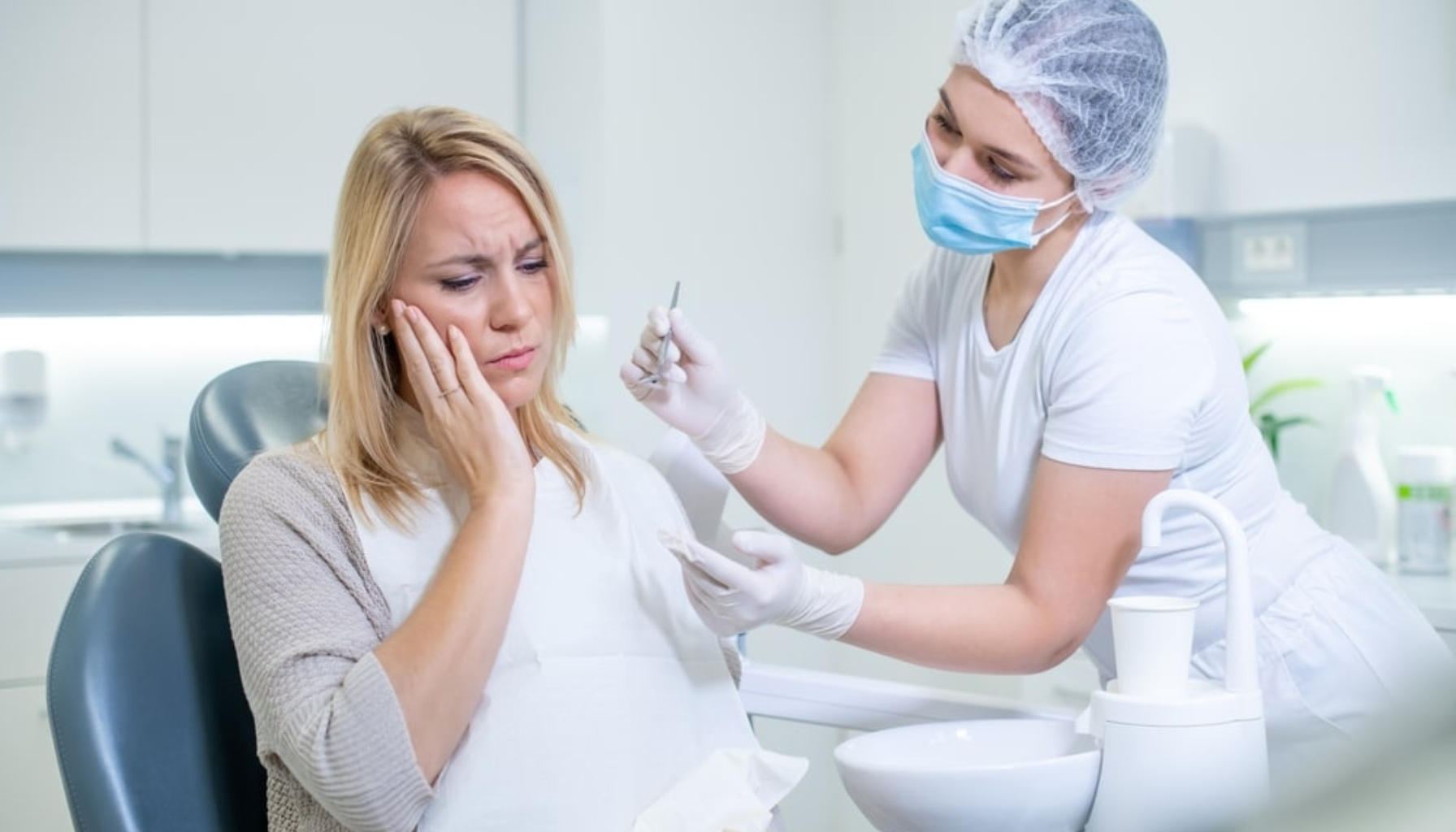 Kvinne med tannverk holder seg til kinnet mens hun forklarer smerten til tannpleier i hvit uniform og munnbind.